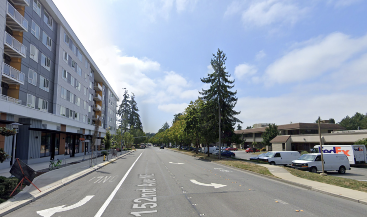 Google Street View image showing one side of the street with a mixed-use building facing the sidewalk, which has a separated bike lane space. The other side has a sharrow, a small sidewalk and then a parking lop with an office park in it.